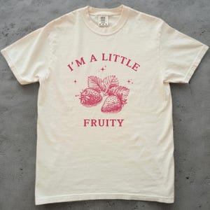 Może przedstawiać: Biały t-shirt z różowym nadrukiem, na którym widnieje napis "I'm a little fruity" oraz rysunek dwóch truskawek i gwiazd.