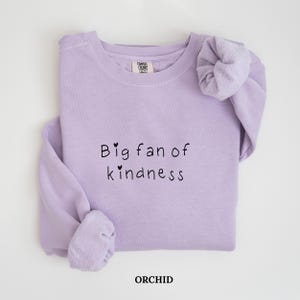 Op de afbeelding: Een lila sweatshirt met de tekst "Big fan of kindness" in zwarte letters. Het woord "ORCHID" is in het zwart gedrukt. De sweatshirt heeft een ronde hals en lange mouwen.