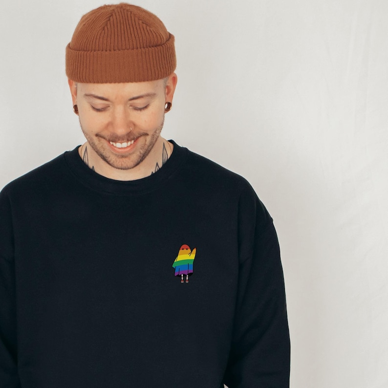 A cute small rainbow ghost waving on the top left pocket of a black sweatshirt