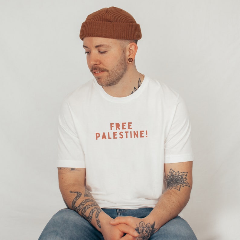 K&ouml;nnte beinhalten: Ein wei&szlig;es T-Shirt mit dem Text "FREE PALESTINE!" in Rot auf der Vorderseite. Die Person, die das Shirt tr&auml;gt, tr&auml;gt eine braune M&uuml;tze und hat T&auml;towierungen auf den Armen.