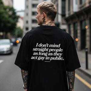 May include: Black oversized t-shirt with white text: "I don't mind straight people as long as they act gay in public." The shirt is worn by a person with blonde hair and tattoos, standing on a city street.
