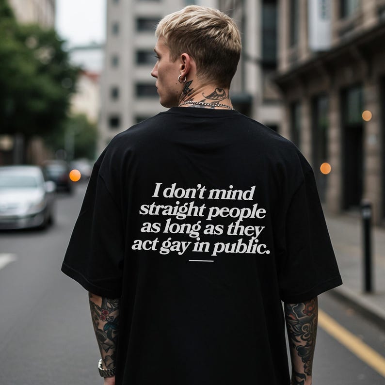 May include: Black oversized t-shirt with white text: "I don't mind straight people as long as they act gay in public." The shirt is worn by a person with blonde hair and tattoos, standing on a city street.