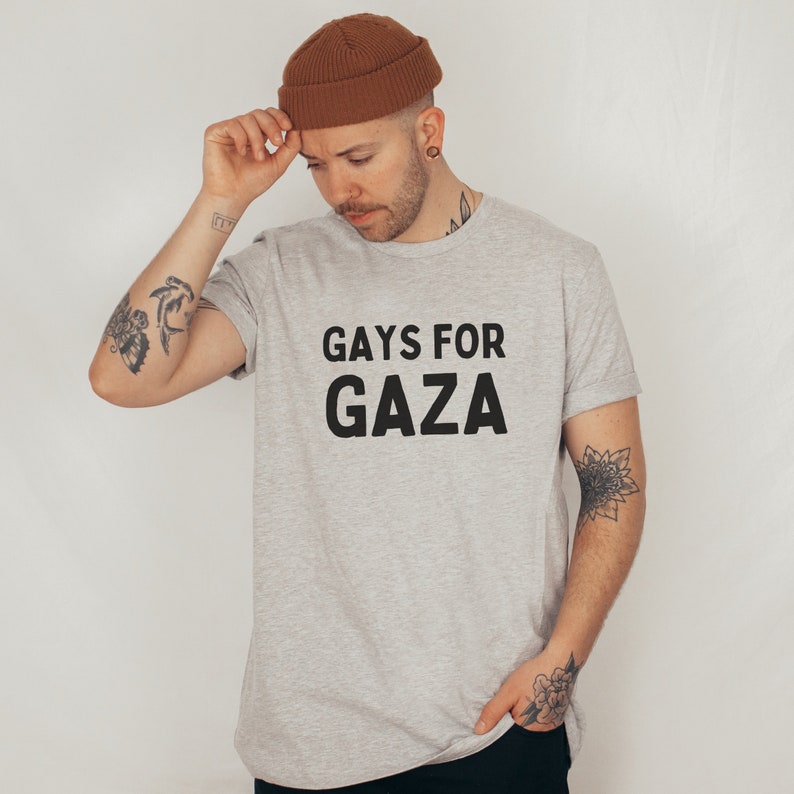 May include: A gray t-shirt with the text "GAYS FOR GAZA" printed in black. The person wearing the shirt is wearing a brown beanie.