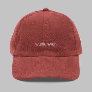 Peut inclure: Casquette de baseball en velours côtelé de couleur rouille avec le mot "wuhluhwuh" brodé en blanc. La casquette a une visière incurvée et une sangle réglable. Un accessoire décontracté.