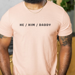 May include: A light pink t-shirt with the text "HE / HIM / DADDY" printed in black.