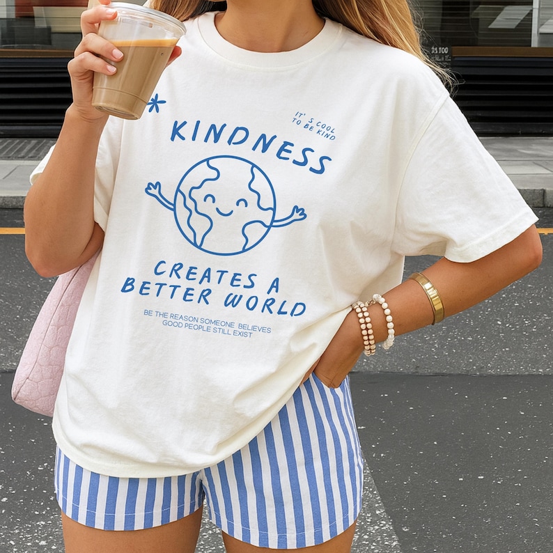 May include: Cream-coloured t-shirt featuring a blue graphic of a smiling globe and the words "KINDNESS CREATES A BETTER WORLD." The shirt also has the text "IT'S COOL TO BE KIND" and "BE THE REASON SOMEONE BELIEVES GOOD PEOPLE STILL EXIST." The model is wearing blue and white striped shorts.