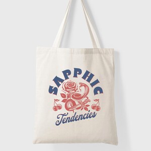 May include: Off-white canvas tote bag with a red and blue graphic. The design includes a rose, snake, cherries, and the words "Sapphic Tendencies". The bag has two long handles for carrying.