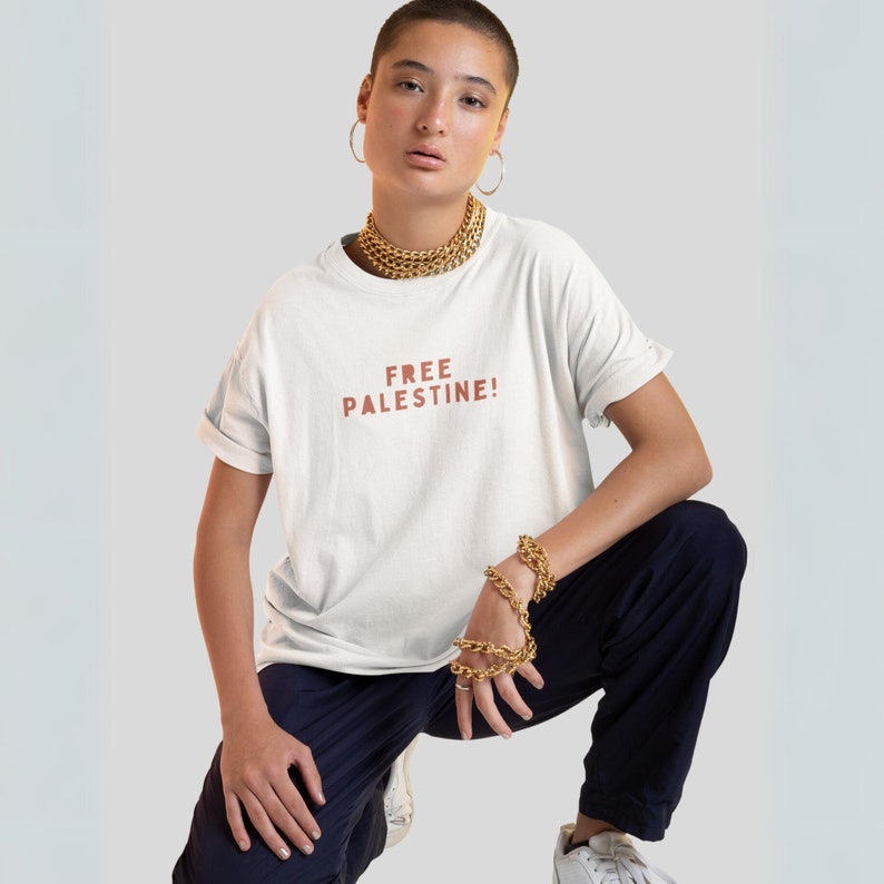 K&ouml;nnte beinhalten: Ein wei&szlig;es T-Shirt mit dem Aufdruck "FREE PALESTINE!" in Braun auf der Vorderseite. Die Person, die das Shirt tr&auml;gt, tr&auml;gt goldenen Schmuck und eine schwarze Hose.
