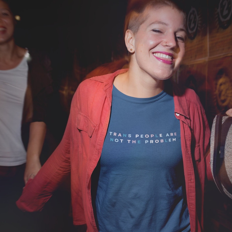 Puede incluir: Una persona lleva una camiseta azul con el texto "TRANS PEOPLE ARE NOT THE PROBLEM" en letras rosa y azul. La persona lleva una camisa roja sobre la camiseta.