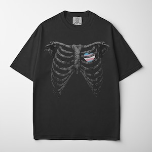 May include: Black t-shirt featuring a detailed illustration of a rib cage with a heart-shaped design in the colors of the transgender flag. The shirt has a crew neck and short sleeves. The tag reads "Comfort Colors XL-10-10".