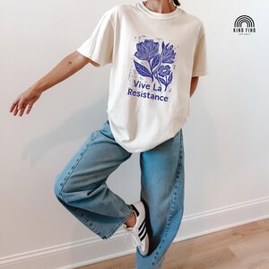 May include: Cream-colored t-shirt with a blue floral design and the text "Vive La Resistance." The shirt is paired with light blue jeans and white sneakers with black stripes. The shirt has short sleeves and a crew neck.
