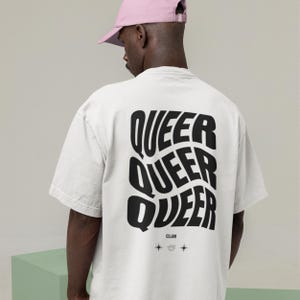 May include: White t-shirt with the word "QUEER" repeated three times in a stacked design on the back. Below the word "QUEER" is the word "CLUB" and two small star and planet graphics. The person in the image is wearing a pink baseball cap.
