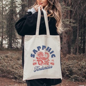 May include: Cream-colored tote bag with the words "Sapphic Tendencies" in blue script. The design includes a red rose, snake, cherries, and other decorative elements. The bag has two cream-colored handles.
