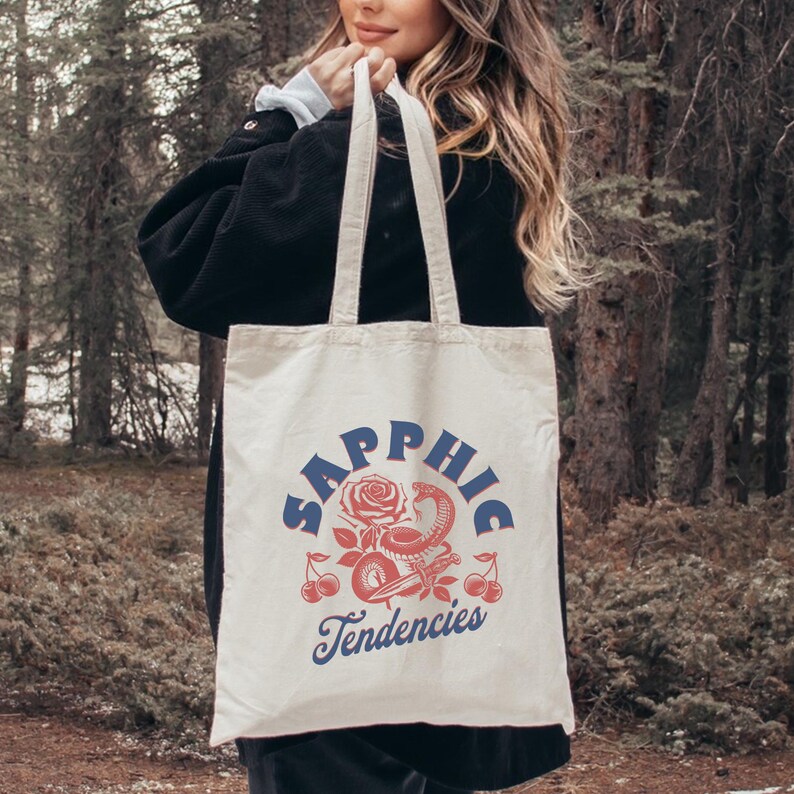 May include: Cream-colored tote bag with the words "Sapphic Tendencies" in blue script. The design includes a red rose, snake, cherries, and other decorative elements. The bag has two cream-colored handles.