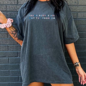 Puede incluir: Una camiseta gris oscuro de gran tama&ntilde;o con el texto "TRANS PEOPLE ARE NOT THE PROBLEM" impreso en azul, rosa y blanco.