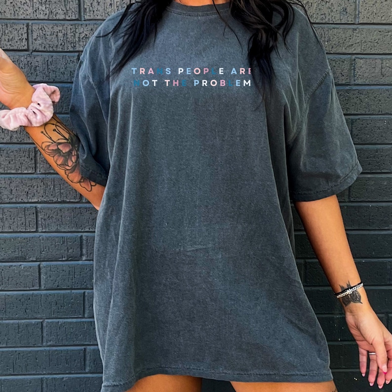 Puede incluir: Una camiseta gris oscuro de gran tama&ntilde;o con el texto "TRANS PEOPLE ARE NOT THE PROBLEM" impreso en azul, rosa y blanco.