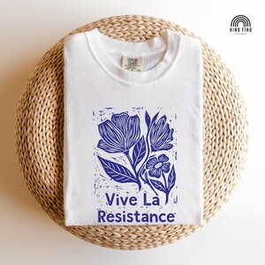 May include: A white crew neck t-shirt featuring a blue block-print style graphic of flowers and leaves. The text "Vive La Resistance" is printed in blue below the floral design. The shirt has a Comfort Colors brand label at the neckline.