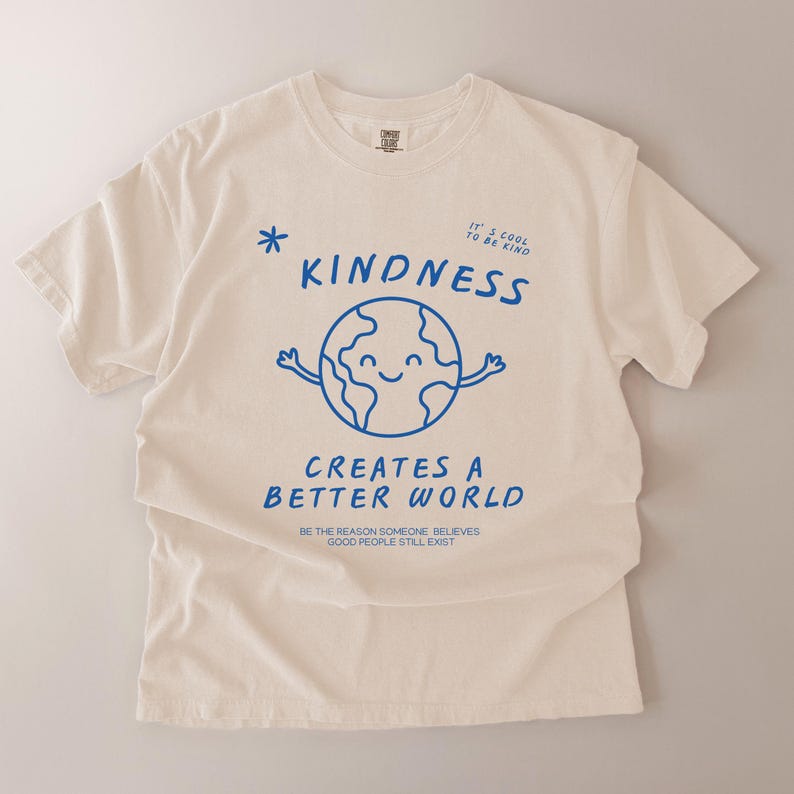 May include: Cream-coloured t-shirt with blue text and graphics. The shirt displays the word "KINDNESS" above a smiling globe with arms. Below, it reads "CREATES A BETTER WORLD" and "BE THE REASON SOMEONE BELIEVES GOOD PEOPLE STILL EXIST."