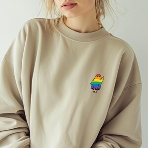 May include: A beige sweatshirt with a small, colourful ghost graphic on the upper left chest. The ghost is designed with rainbow colours. The sweatshirt has a crew neck and long sleeves.
