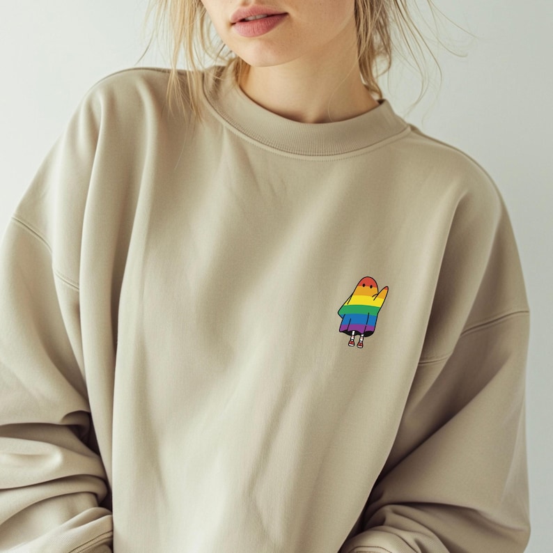 May include: A beige sweatshirt with a small, colourful ghost graphic on the upper left chest. The ghost is designed with rainbow colours. The sweatshirt has a crew neck and long sleeves.
