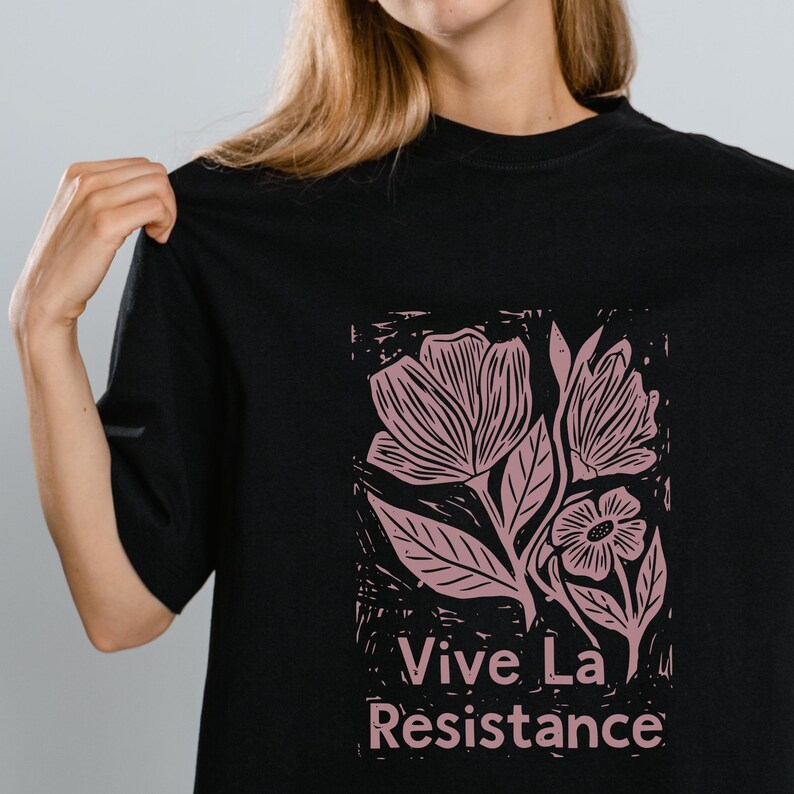 May include: Black t-shirt featuring a pink floral design and the words "Vive La Resistance" in white. The graphic has a woodcut style, with detailed lines and shading. The shirt is a classic crew neck style.