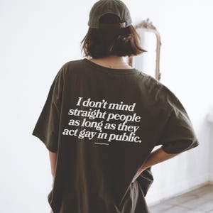 May include: Dark olive green t-shirt with the text "I don't mind straight people as long as they act gay in public" in white. The person is wearing a dark green baseball cap. The shirt is a casual, oversized fit.