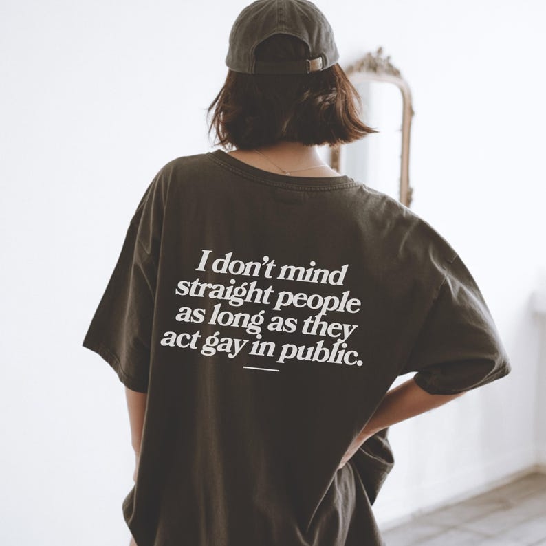May include: Dark olive green t-shirt with the text "I don't mind straight people as long as they act gay in public" in white. The person is wearing a dark green baseball cap. The shirt is a casual, oversized fit.