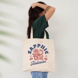 May include: Cream-colored tote bag with the word "Sapphic" in blue arched text above a graphic of a rose, snake, and cherries in red and white. The text "Tendencies" is below the graphic in blue.
