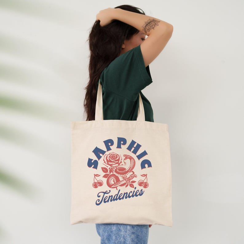 May include: Cream-colored tote bag with the word "Sapphic" in blue arched text above a graphic of a rose, snake, and cherries in red and white. The text "Tendencies" is below the graphic in blue.