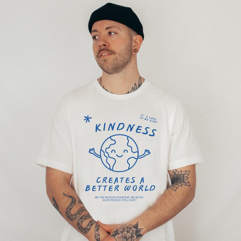 May include: White t-shirt featuring a blue graphic of a smiling globe with arms and the word "KINDNESS". The text "CREATES A BETTER WORLD" is below the globe. The shirt also has the text "BE THE REASON SOMEONE BELIEVES GOOD PEOPLE STILL EXIST".