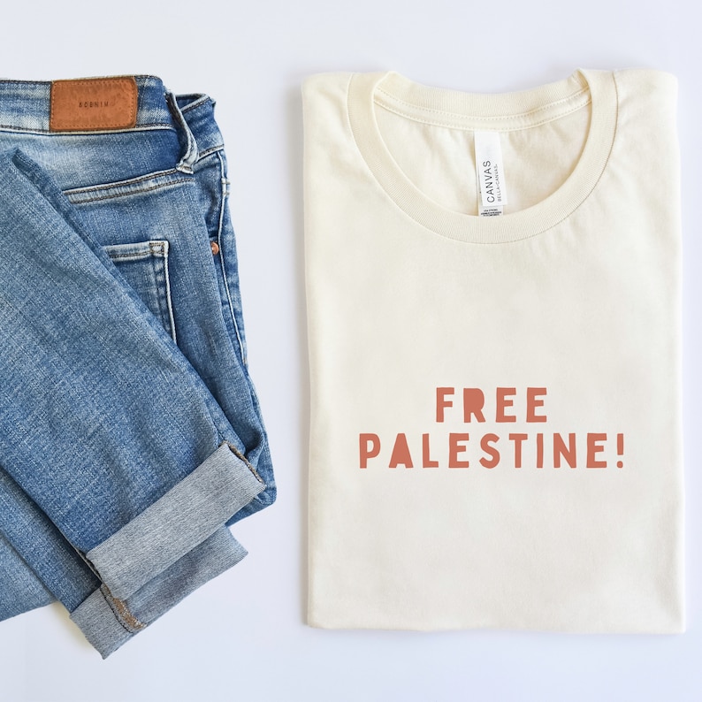 K&ouml;nnte beinhalten: Ein cremefarbenes T-Shirt mit dem Aufdruck "FREE PALESTINE!" in Orange auf der Vorderseite. Das T-Shirt ist gefaltet und liegt flach auf einer wei&szlig;en Oberfl&auml;che. Eine blaue Jeans ist gefaltet und liegt flach auf der linken Seite des Bildes.
