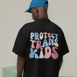 Może przedstawiać: Czarny T-shirt z napisem "PROTECT TRANS KIDS" w kolorowych, stylizowanych literach.