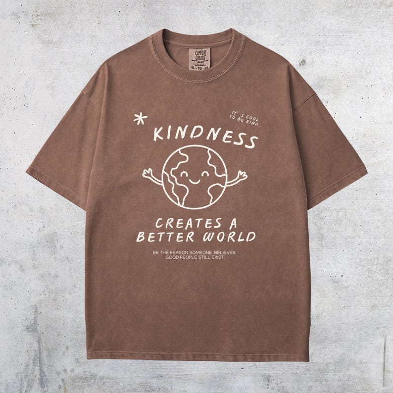 May include: Brown t-shirt with the word "KINDNESS" and a smiling earth graphic. The shirt also features the text "CREATES A BETTER WORLD" and "IT'S COOL TO BE KIND". The shirt is a Comfort Colors brand.