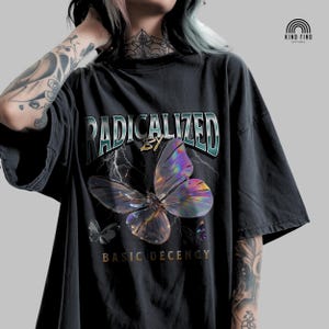 Könnte beinhalten: Schwarzes T-Shirt mit dem Text "RADICALIZED BY BASIC DECENCY" in Türkis und Gold. Eine große, schillernde Schmetterlingsgrafik ist zentral platziert, mit kleineren Schmetterlingsillustrationen. Das T-Shirt hat einen Vintage-Look.