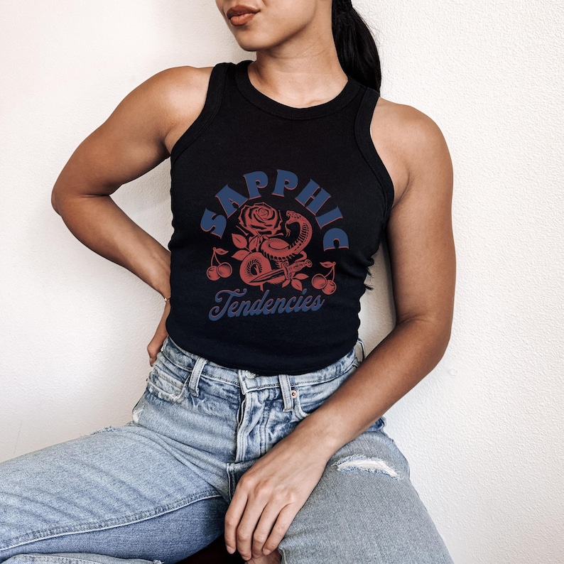 Puede incluir: Camiseta sin mangas negra con un dise&ntilde;o gr&aacute;fico que presenta el texto "Sapphic Tendencies" en azul y una ilustraci&oacute;n de rosa roja, serpiente y cerezas. La camiseta sin mangas es de estilo acanalado.