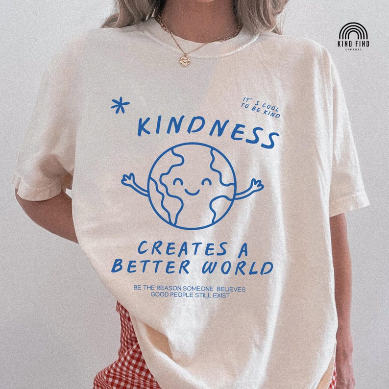 May include: Cream-coloured t-shirt featuring a blue graphic of a smiling globe with arms, the word "KINDNESS", and the phrase "CREATES A BETTER WORLD". The shirt also has the text "IT'S COOL TO BE KIND" and "BE THE REASON SOMEONE BELIEVES GOOD PEOPLE STILL EXIST".