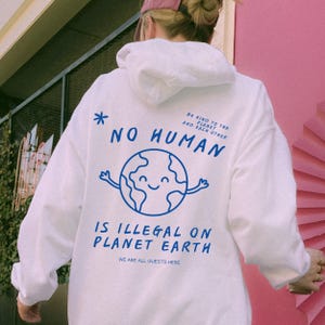 Puede incluir: Sudadera blanca con texto azul y un gráfico de la Tierra sonriente. El texto dice "NO HUMAN IS ILLEGAL ON PLANET EARTH" con la frase "WE ARE ALL GUESTS HERE" debajo. La sudadera también tiene el texto "BE KIND TO THE PLANET AND EACH OTHER".