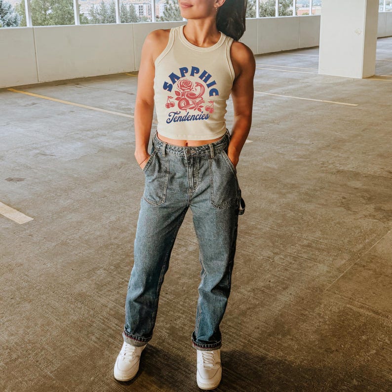 Puede incluir: Camiseta sin mangas color crema con el texto "Sapphic Tendencies" en azul y rosa, con un dise&ntilde;o de rosa y cerezas. Se combina con vaqueros azules y zapatillas blancas. El conjunto es informal y elegante.