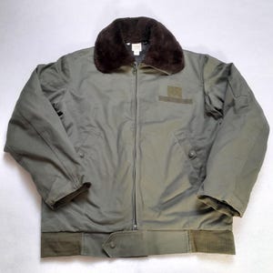 Peut inclure: Un blouson vert olive doté d'un col en fausse fourrure marron foncé, d'une fermeture éclair intégrale, de deux poches latérales à boutons-pression et d'un ourlet côtelé. Un patch auto-agrippant rectangulaire est situé sur la poitrine gauche.