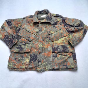 Può includere: Giacca mimetica con un motivo screziato marrone, verde e nero. La giacca ha un collo, più tasche frontali e chiusure con bottoni. Le maniche sono lunghe e la giacca sembra essere realizzata in un tessuto resistente.