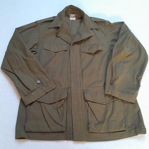 Peut inclure: Une veste de terrain vert olive dotée d'un col pointu, de manches longues avec poignets boutonnés et de quatre grandes poches avant à rabats boutonnés. La veste comprend des épaulettes et une patte de boutonnage centrale avec une étiquette.