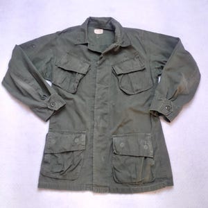 Könnte beinhalten: Olivgrüne Jacke im Militärstil mit vier Vordertaschen und geknöpften Manschetten. Die Jacke hat einen Kragen und eine Knopfleiste. Der Stoff scheint abgenutzt zu sein, was auf einen Vintage-Zustand hindeutet. Die Jacke liegt flach auf einer hellen Oberfläche.