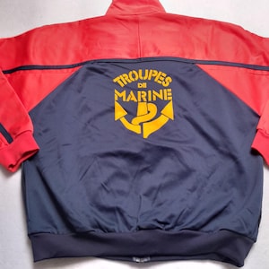 May include: A vintage red and navy blue jacket featuring the text "TROUPES DE MARINE" and a yellow anchor emblem on the back. The jacket has a stand-up collar and red cuffs.