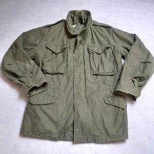 May include: An olive green military-style jacket with four front pockets and a button-up closure. The jacket has a collared neckline and long sleeves with buttoned cuffs. The fabric appears to be a durable, possibly water-resistant material.