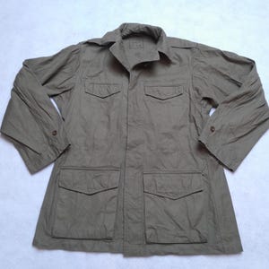 Puede incluir: Chaqueta verde oliva con cuatro bolsillos delanteros, cuello y puños abotonados. La chaqueta tiene un corte holgado y parece estar hecha de una tela ligera. Tiene cierre de botones y una pequeña etiqueta cerca del cuello.