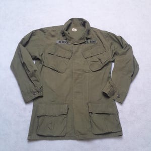 Könnte beinhalten: Olivgrüne Militärjacke mit vier Vordertaschen und zwei Brusttaschen. Die Jacke hat einen Kragen und geknöpfte Manschetten. Der Name "RENSEL" und "U.S. ARMY" sind auf der Jacke zu sehen.