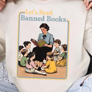 Op de afbeelding: Een wit sweatshirt met de tekst "Let's Read Banned Books" in een retro lettertype. Het ontwerp toont een leraar die een boek voorleest aan een groep kinderen.