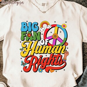 Könnte beinhalten: Cremefarbenes T-Shirt mit dem Text "BIG FAN OF HUMAN RIGHTS" in einem Retro-Schriftzug. Das Design umfasst ein Friedenszeichen, einen Regenbogen, Blumen und eine geballte Faust. Rundhalsausschnitt.