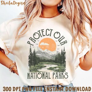 Peut inclure: Un t-shirt blanc avec un motif graphique de style vintage qui dit "Prot&eacute;gez nos parcs nationaux". Le motif repr&eacute;sente une cha&icirc;ne de montagnes, un coucher de soleil et une rivi&egrave;re.