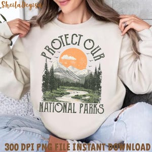 Peut inclure: Un sweat-shirt blanc avec un motif graphique de style vintage repr&eacute;sentant une cha&icirc;ne de montagnes, un coucher de soleil et une rivi&egrave;re. Le texte "Protect Our National Parks" est imprim&eacute; en marron sur le sweat-shirt.
